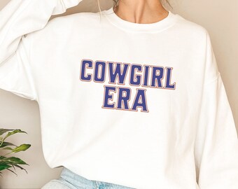 May include: White long-sleeved sweatshirt with the text "COWGIRL ERA" in blue and orange lettering.