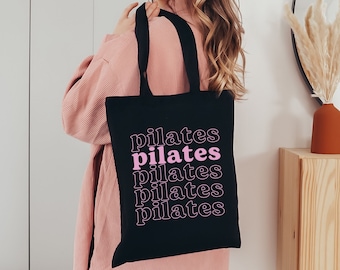 May include: A black tote bag with the word "pilates" repeated four times in pink lettering. The bag is being held by a person wearing a pink sweater.