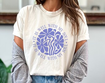 May include: Ivory-colored t-shirt featuring a circular design with the text "IT IS WELL WITH MY SOUL" in a light purple hue. The design includes stylized floral elements in the same color. The shirt is a casual, comfortable style.