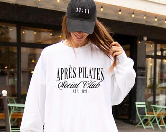 May include: A white long-sleeve sweatshirt featuring the black text APRÈS PILATES Social Club - EST. 2025 - on the chest, paired with a black baseball cap embroidered with the white numbers 11:11 on the front panel.