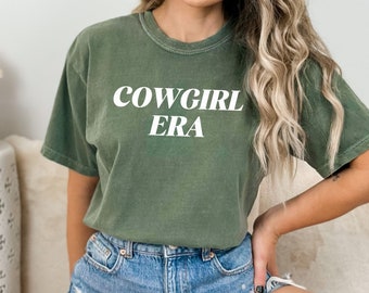 May include: A green t-shirt with the text "COWGIRL ERA" printed in white on the front. The shirt is worn by a person with long blonde hair and is paired with denim shorts.