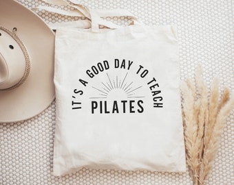 May include: White canvas tote bag with black text that reads "It's a good day to teach Pilates". The text is in a semi-circle shape with a sunburst design in the center.