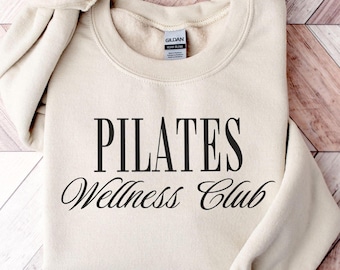 May include: A cream-colored Pilates Wellness Club sweatshirt with black text. The sweatshirt is folded on a light brown wooden surface. The Gildan brand tag is visible on the inside of the collar.