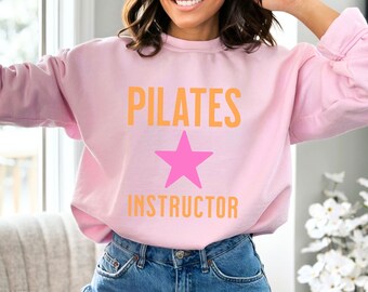 May include: A light pink long-sleeve crewneck sweatshirt featuring the orange text PILATES INSTRUCTOR with a bright pink five-pointed star graphic positioned between the two words. The garment has a ribbed collar, cuffs, and hemline.