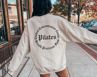 Può includere: Una felpa color crema con una grafica circolare nera sul retro. La grafica dice "Pilates" in un font grande e "Control Concentration Center Breath Precision Flow" in un font più piccolo intorno all'esterno del cerchio.
