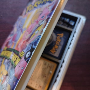 May include: A white plastic case with a Pokemon VHS tape inside. The case is open and the tape is visible. The tape has a colorful image of Pokemon characters on it. The text on the tape reads "Pokemon: The Movie 2000".