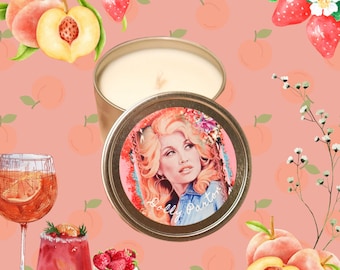 Puede incluir: Una lata de vela dorada con un retrato de Dolly Parton en la tapa. La imagen está ambientada sobre un fondo color melocotón con ilustraciones de acuarela de melocotones, fresas y cócteles. Un artículo decorativo.