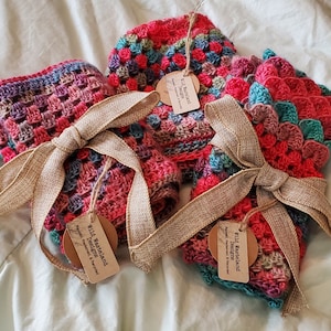 May include: Three crocheted hand warmers in shades of pink, red, and teal with a brown and white ribbon tied around them. The hand warmers are labeled "Wild Wasteland Designs" and "Reclaimed & Recycled".