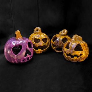 Può includere: Quattro zucche di Halloween in ceramica con finiture lucide. Una è viola con un ritaglio a forma di cuore, e le altre sono arancioni con facce intagliate. Le zucche hanno gambi e sono su uno sfondo nero.