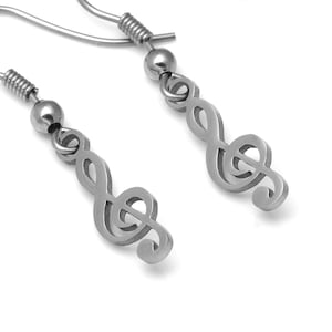 Könnte beinhalten: Ein Paar silberfarbene Ohrringe mit einem Notenschlüssel-Design. Jeder Ohrring hat einen Haken, eine kleine Kugel und ein stilisiertes Musiksymbol. Die Ohrringe werden vor einem weißen Hintergrund präsentiert.