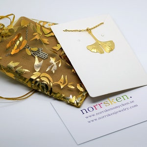 May include: Gold ginkgo leaf necklace on a white card with the text "norrsken. www.norrskensmycken.se www.norrskenjewelry.com"