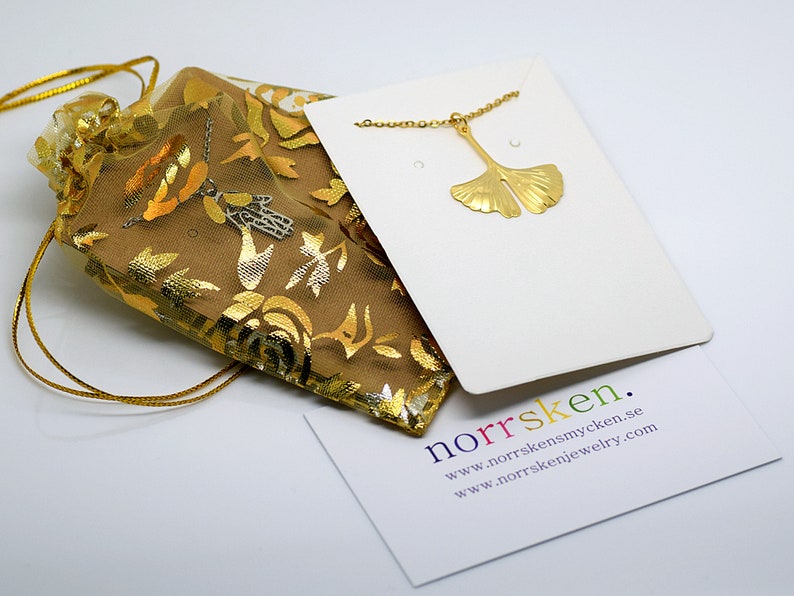 May include: Gold ginkgo leaf necklace on a white card with the text "norrsken. www.norrskensmycken.se www.norrskenjewelry.com"