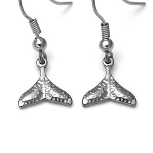 May include: A pair of silver-colored earrings. Each earring features a fish tail charm suspended from a hook. The tail charm has a textured surface and is detailed with small indentations along the bottom edge.