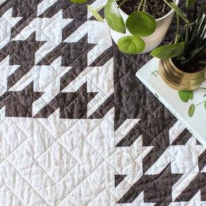 Pode incluir: Um edredom branco e cinza com um padrão de pata de galo. O edredom está sobre uma mesa com duas plantas em vasos e um livro intitulado "The New Plant Parent".