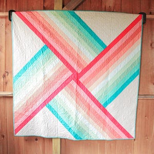 May include: A colorful quilt with a geometric design. The quilt features diagonal stripes in shades of red, coral, turquoise, and green on a white background. The quilt is hanging on a wooden wall.