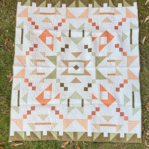 May include: A white quilt with a geometric pattern featuring orange, green, and brown shapes. The quilt has a border of green triangles and is laid out on a grassy surface. The design includes squares, triangles, and diamond shapes.