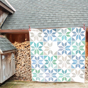 May include: A large, colorful quilt with a geometric starburst pattern in shades of blue, green, and gray against a white background. The quilt is hanging outside on a wooden structure, showcasing its design and craftsmanship.