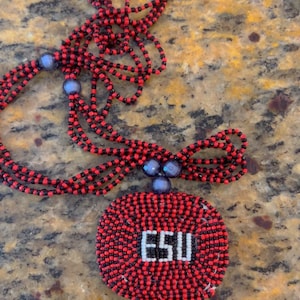 Puede incluir: Un collar de cuentas rojas y negras con un colgante redondo que presenta las letras "ESU" en blanco. El colgante est&aacute; rodeado por un borde de cuentas negras y rojas.