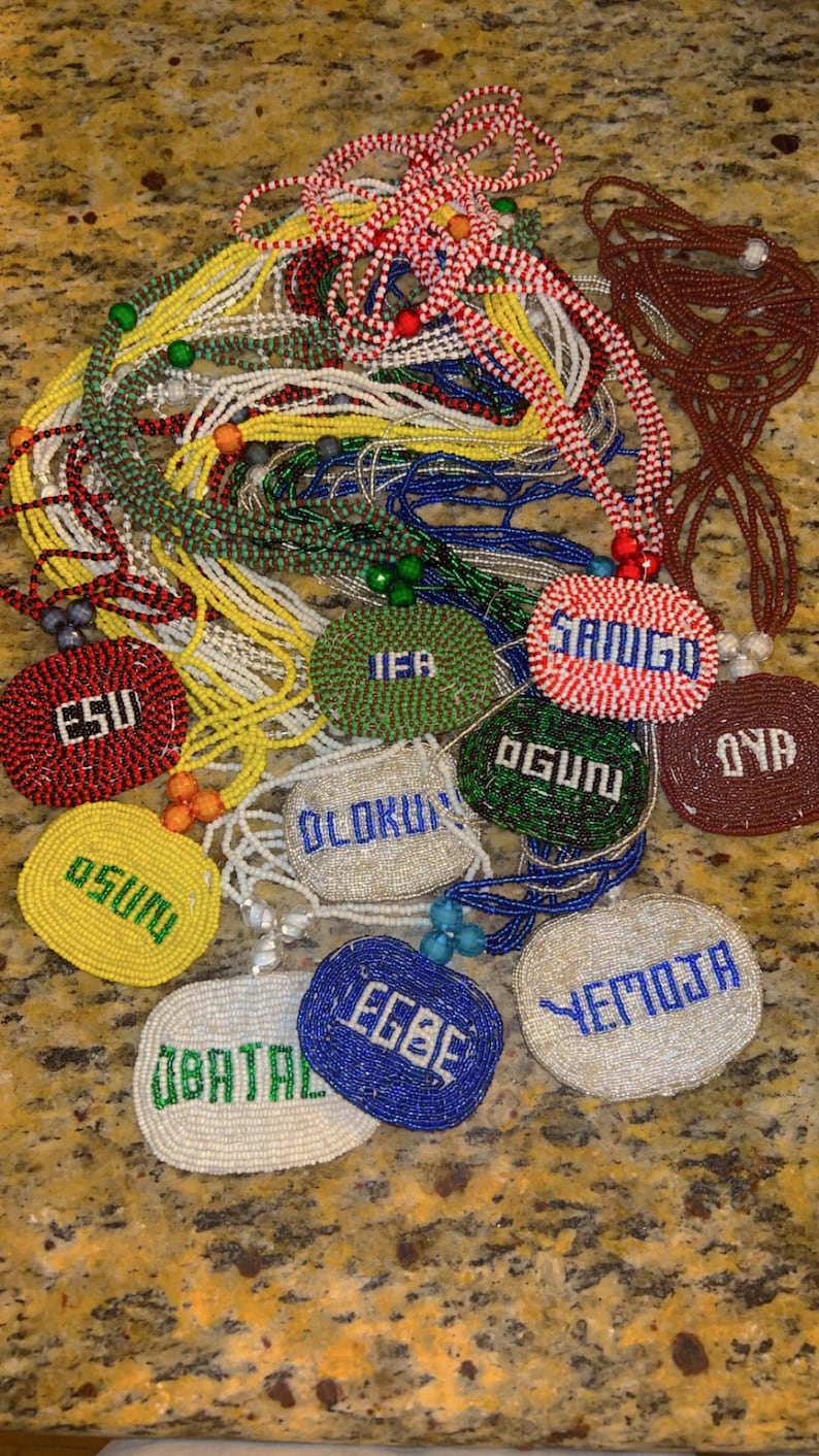 Puede incluir: Una colecci&oacute;n de collares de cuentas de colores con colgantes de cuentas de forma ovalada. Los colgantes tienen diferentes nombres escritos en ellos, incluyendo "Esu", "Osun", "Olokun", "Ifa", "Ogun", "Yemoja", "Egbere", "Obatala", "Oya" y "Sango".