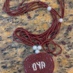Puede incluir: Un collar de cuentas marrones con un colgante circular que dice "OYA" en cuentas blancas.