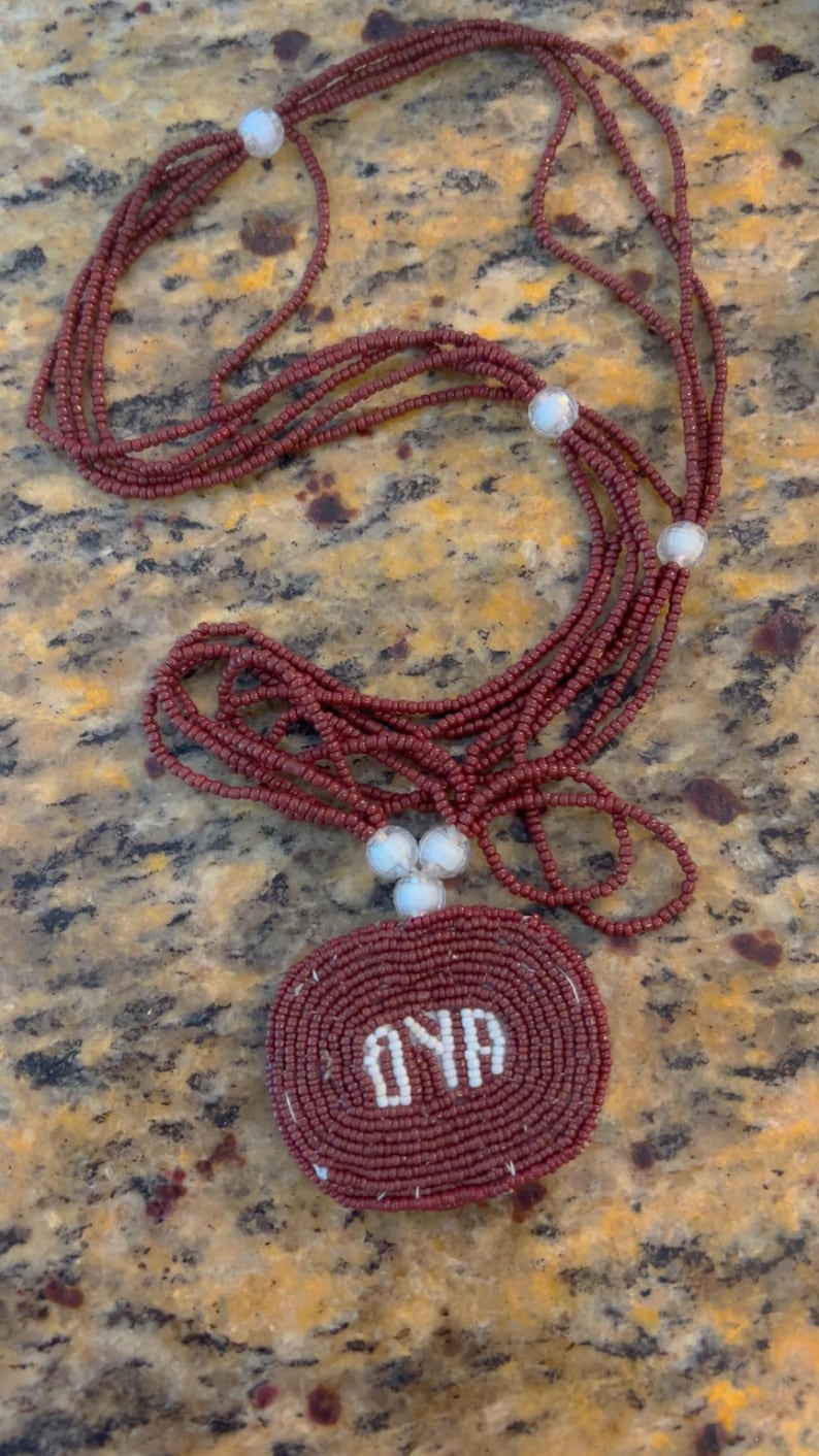 Puede incluir: Un collar de cuentas marrones con un colgante circular que dice "OYA" en cuentas blancas.