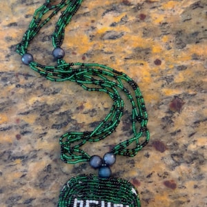 Puede incluir: Un collar de cuentas verdes y negras con un colgante redondo que dice "OGUN" en cuentas blancas.