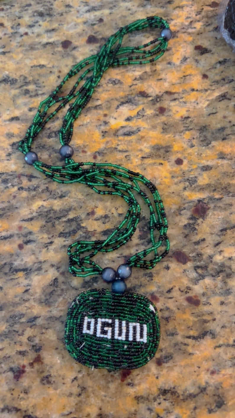 Puede incluir: Un collar de cuentas verdes y negras con un colgante redondo que dice "OGUN" en cuentas blancas.