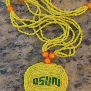 Puede incluir: Un collar de cuentas amarillas con un acento de cuentas naranjas y un colgante redondo de cuentas amarillas con la palabra "OSU" en cuentas verdes.
