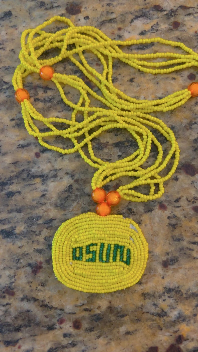 Puede incluir: Un collar de cuentas amarillas con un acento de cuentas naranjas y un colgante redondo de cuentas amarillas con la palabra "OSU" en cuentas verdes.