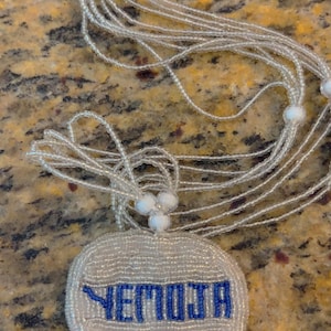 Puede incluir: Un collar de cuentas blancas con un colgante redondo que dice "YETMOJA" en cuentas azules.