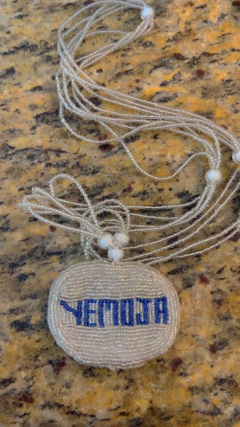 Puede incluir: Un collar de cuentas blancas con un colgante redondo que dice "YETMOJA" en cuentas azules.