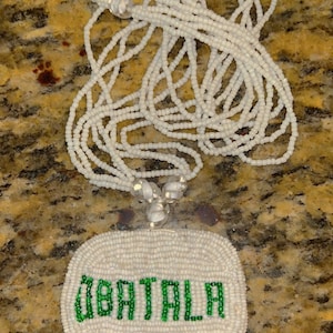 Puede incluir: Collar de cuentas blancas con un colgante ovalado blanco que dice "OBATALA" en cuentas verdes.