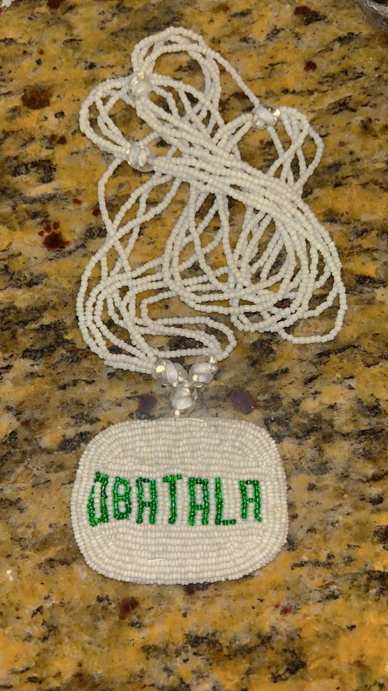 Puede incluir: Collar de cuentas blancas con un colgante ovalado blanco que dice "OBATALA" en cuentas verdes.