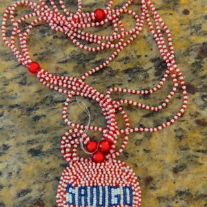 Puede incluir: Un collar de cuentas rojas y blancas con un colgante redondo que dice "SAUVOIS" en cuentas azules y blancas.