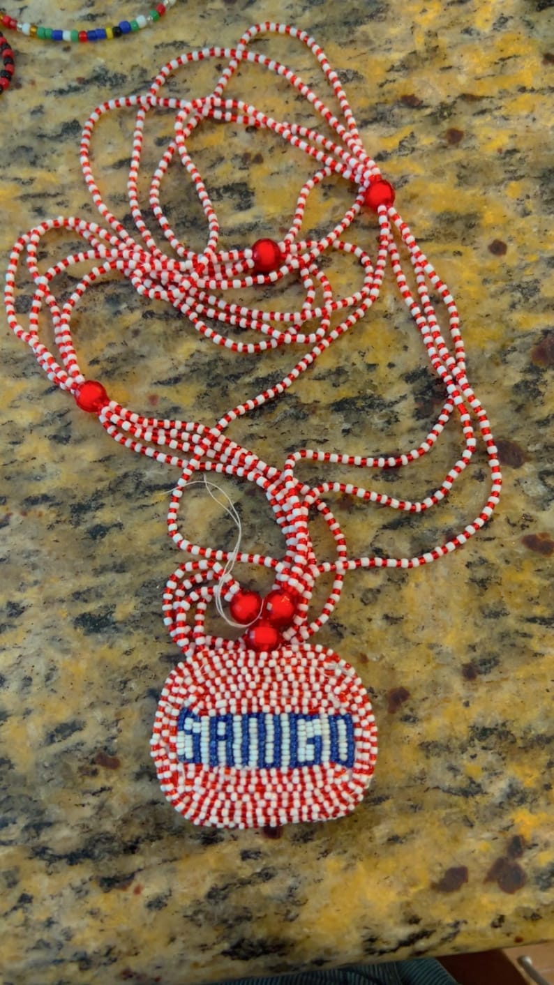 Puede incluir: Un collar de cuentas rojas y blancas con un colgante redondo que dice "SAUVOIS" en cuentas azules y blancas.