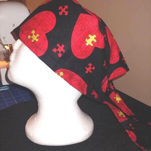 May include: A black and red patterned head covering with a puzzle heart design. The fabric features large red puzzle piece hearts and gold puzzle pieces. The head covering is displayed on a white mannequin head.