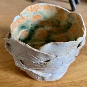May include: A handmade ceramic bowl with a textured, leaf-like exterior in shades of white and light blue. The interior features orange and green accents. This unique piece is ideal for serving or display.