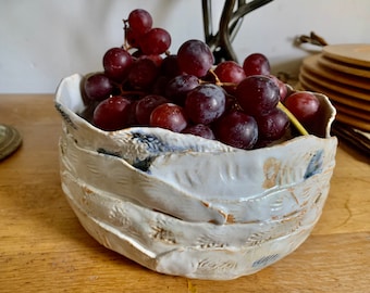 May include: A white ceramic bowl with a textured, layered design, filled with ripe red grapes. The bowl has blue and brown accents and sits on a wooden surface. A decorative piece for the kitchen or dining room.