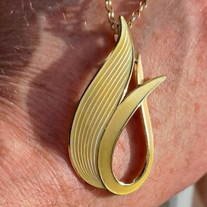 Peut inclure: Un collier avec un pendentif en or en forme de flamme stylisée. Le pendentif a une surface texturée et une forme incurvée lisse.