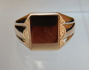 Könnte beinhalten: Ein goldener Siegelring mit rechteckiger Oberfläche. Der Ring hat eine polierte Oberfläche und dekorative Details an den Schultern. Die Oberfläche des Rings ist dunkel und reflektierend.