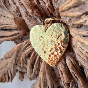 May include: A gold heart-shaped pendant with a textured surface. The pendant hangs from a small gold loop. The background is a piece of weathered, light brown wood.