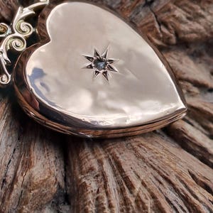 Può includere: Un medaglione a forma di cuore, color oro, con un motivo a stella. Il medaglione ha un'apertura a cerniera e un'ansa decorativa. Il medaglione è appoggiato su una superficie di legno marrone strutturata. Gioiello vintage.