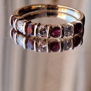 May include: A gold band ring with alternating red and clear gemstones. The ring showcases a series of vibrant red stones and sparkling clear stones, creating a striking contrast. The ring is displayed on a reflective surface.