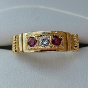 Puede incluir: Un anillo de banda de oro con tres piedras engastadas en un marco rectangular. La piedra central es un diamante claro, y las dos piedras exteriores son rubíes rojos.