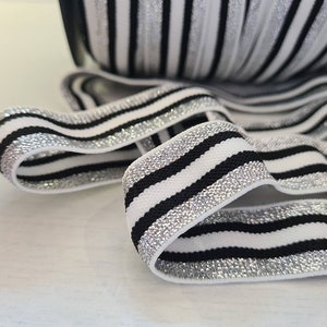 Puede incluir: Elástico a rayas blancas y negras con purpurina plateada. El elástico está doblado y muestra el patrón.