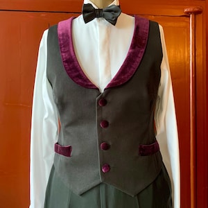 Pode incluir: Um conjunto formal com um colete preto com acabamento em veludo roxo escuro e botões a condizer. É combinado com uma camisa branca, uma gravata borboleta preta e calças pretas. A roupa é exibida num manequim.