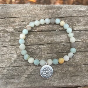 Puede incluir: Un brazalete de cuentas de color verde claro y gris con un dije de plata con dise&ntilde;o de flor de loto.