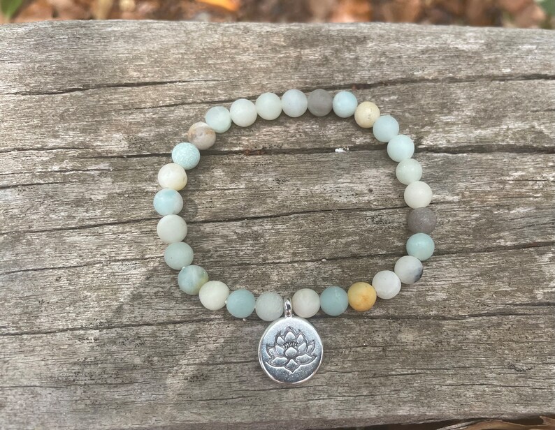 Puede incluir: Un brazalete de cuentas de color verde claro y gris con un dije de plata con dise&ntilde;o de flor de loto.