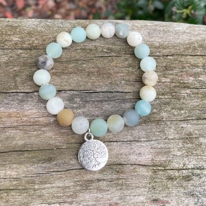 Puede incluir: Un brazalete de cuentas verdes, azules y marrones con un dije de plata de &aacute;rbol de la vida.