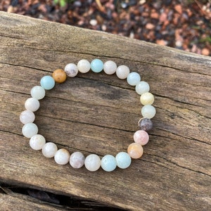 Puede incluir: Un brazalete hecho de cuentas de color azul claro, blanco y rosa. Las cuentas son redondas y tienen una forma natural ligeramente irregular. El brazalete est&aacute; sobre una superficie de madera.